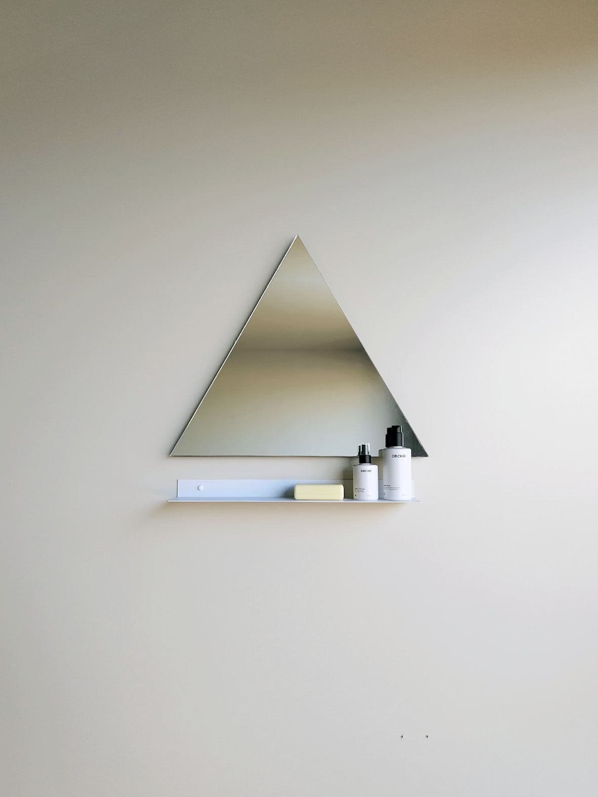 A steel shelf below a triangle bathroom mirror. The modern bathroom shelf holds an artisan soap and two soap dispensers from Mr Price home. The wall mirror and shelf are in a modern bathroom design in Johannesburg