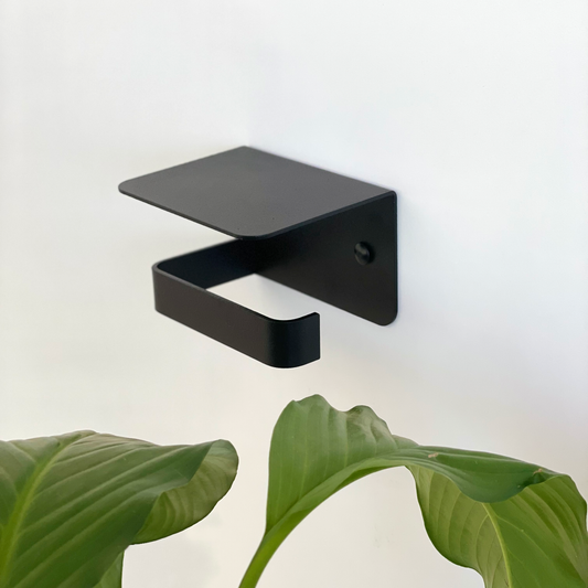 A black steel toilet paper holder or black steel toilet roll holder with shelf in a Tile Africa store with an indoor bathroom plant from Mr Price home