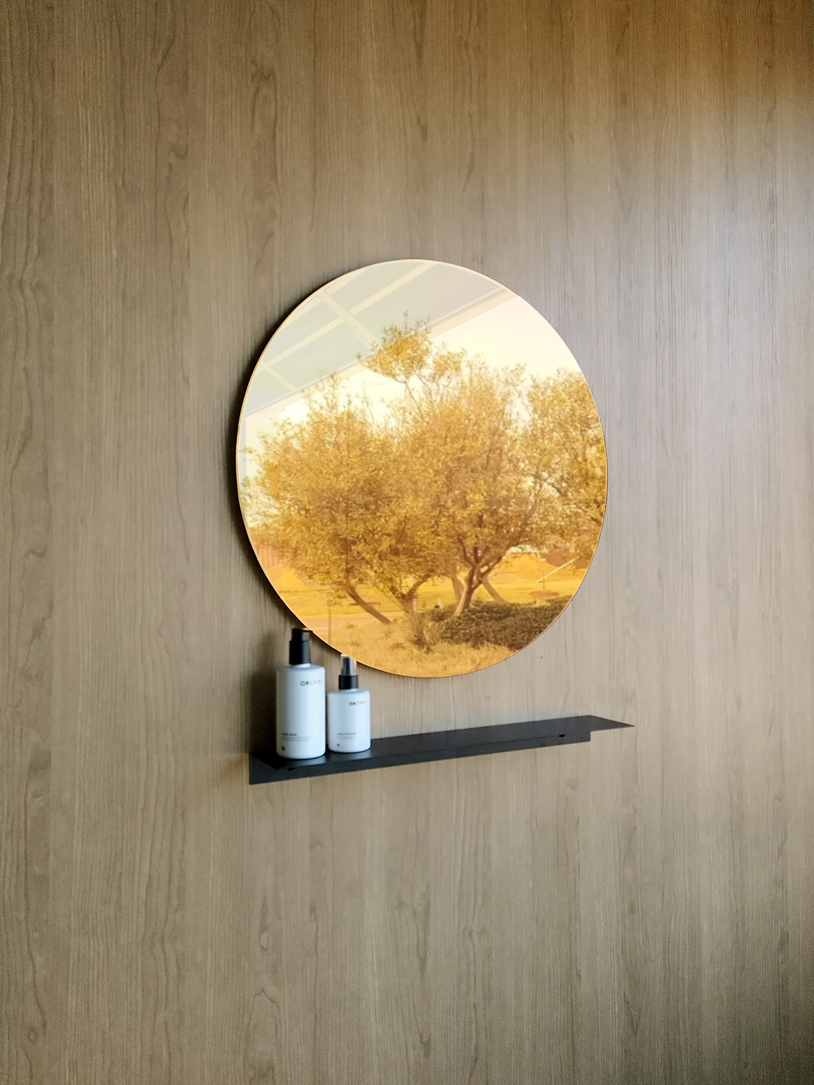 A big beautiful round wall mirror in a luxury bathroom design in Johannesburg. The large round mirror has a reflection of nature. The gold round mirror hangs above a black steel shelf. The shelf for bathroom holds two soap dispensers from Mr Price Home