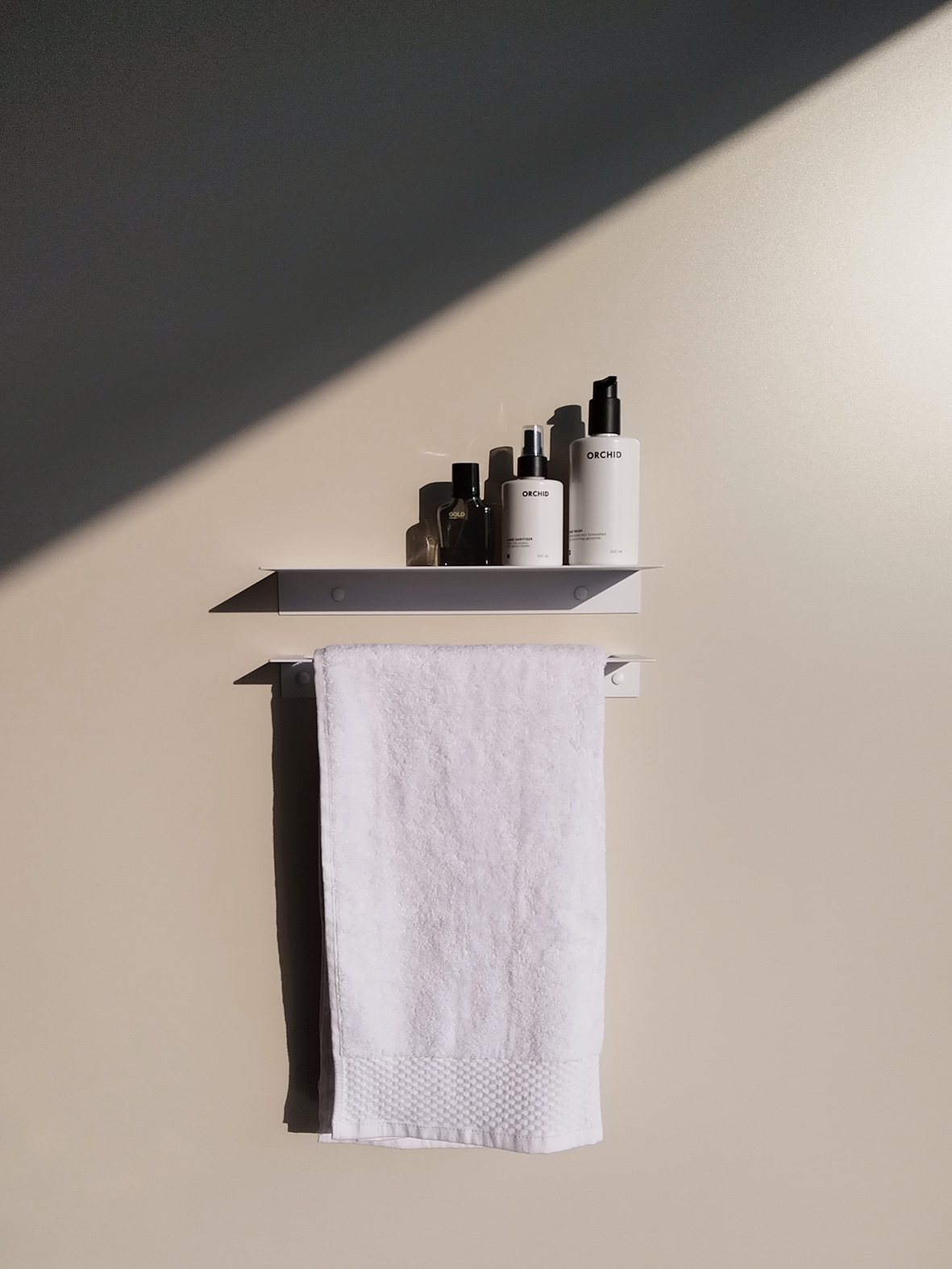 A metal bathroom shelf in a modern bathroom design in Johannesburg. The metal shelf has a cologne bottle and two soap dispenser bottles from Mr Price home. Below the modern bathroom shelf is a steel towel rack that holds a white towel